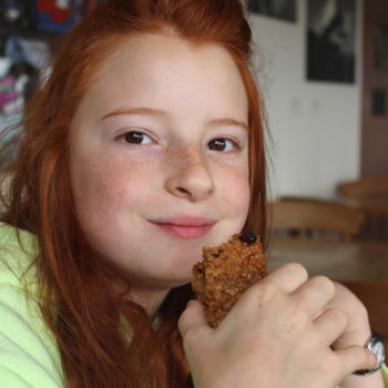 Photo showing a young girl eating a rectangular homemade flapjack biscuit made with oats, raisins, cherries and golden syrup.  The flapjack biscuit is pictured in her hands and she takes a big bite from the end of the biscuit.